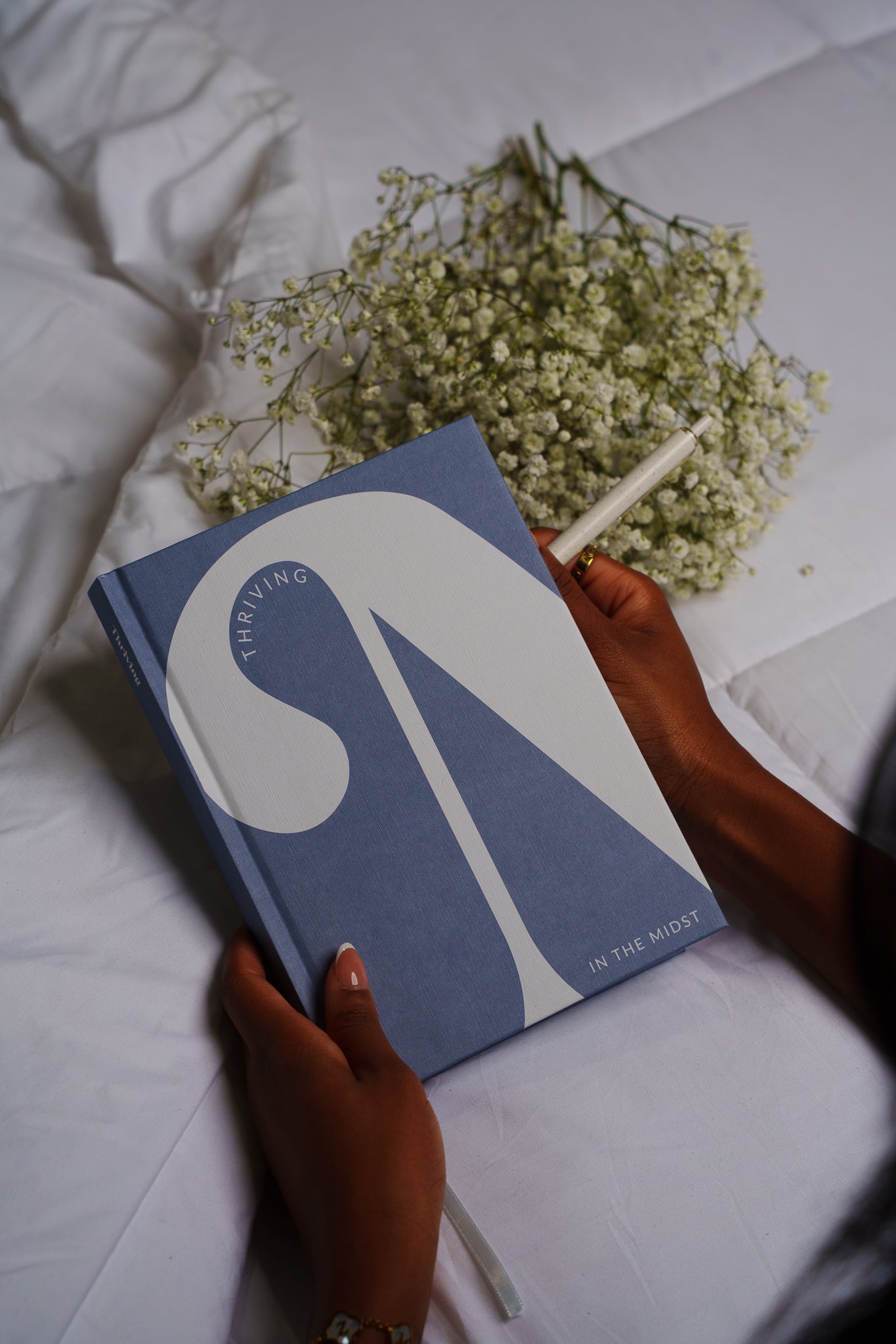 Blue book with a white abstract design held by a hand on a white surface with flowers.